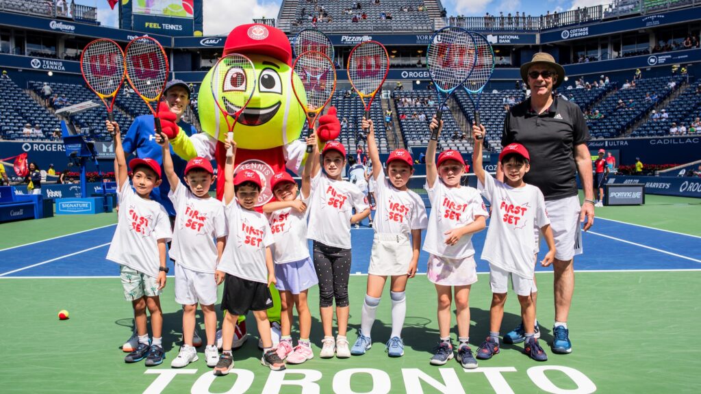 A group of children in the Rogers First Set program stand on Centre Court with the tournament mascot holding racquets in the air.