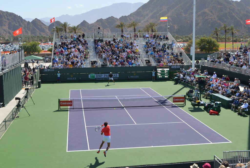 Wide shot of Gabriel Diallo (close side with back to camera) hitting a forehand in Indian Wells with the mountains in the background.