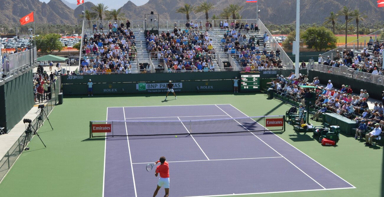 Wide shot of Gabriel Diallo (close side with back to camera) hitting a forehand in Indian Wells with the mountains in the background.